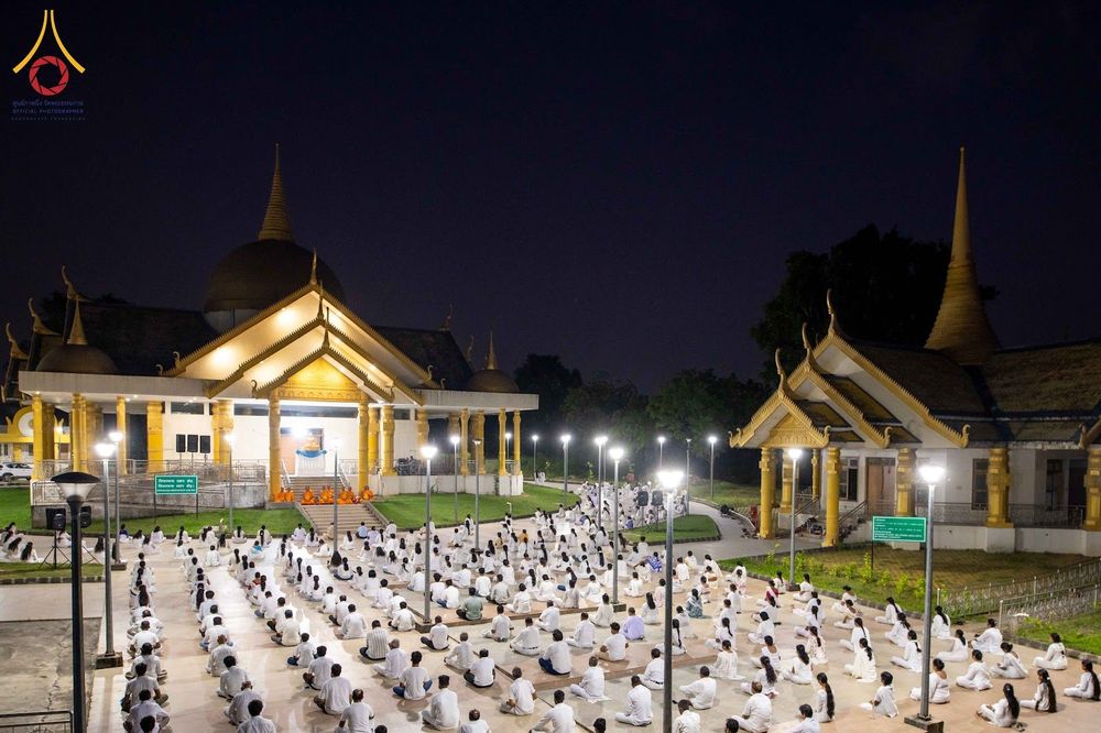 ผู้คนสวมชุดขาวนั่งสมาธิบนลานกว้างหน้าอาคารทรงไทยในยามค่ำคืน
