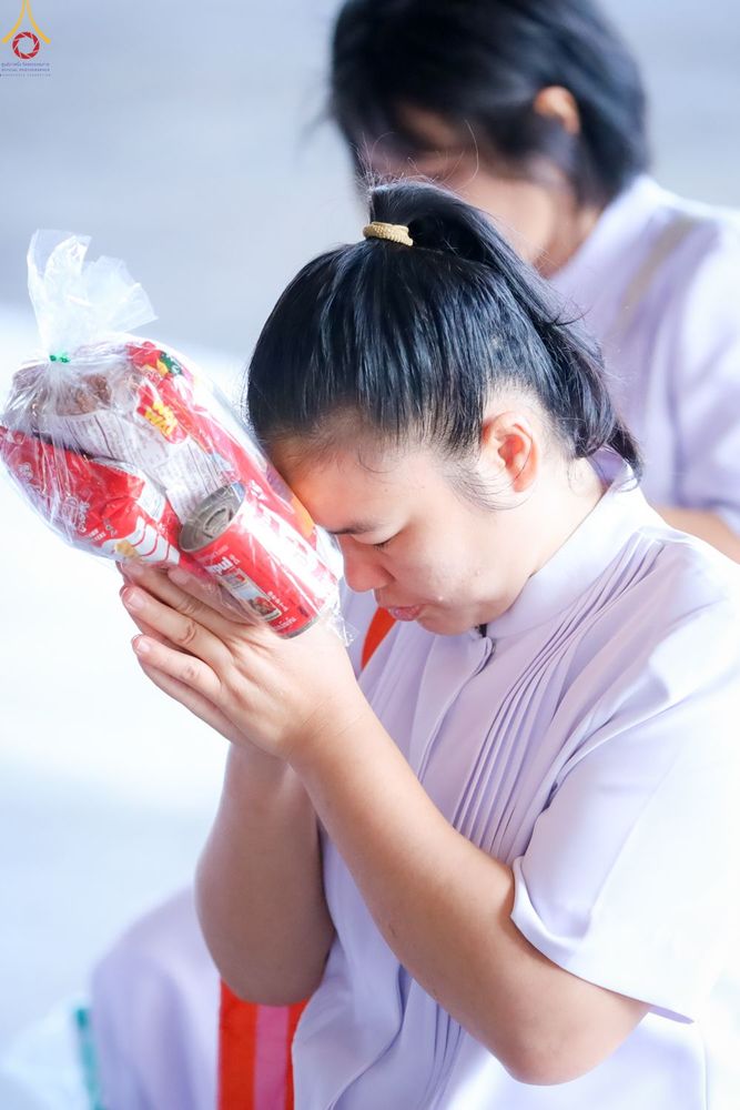 หญิงสาวสวมชุดสีขาวกำลังพนมมือถือถุงใส่ของตักบาตรด้วยท่าทางสงบและตั้งใจ
