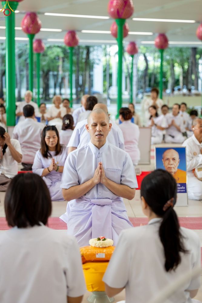 ผู้ชายในชุดสีขาวนั่งพนมมือในพิธีทางศาสนาท่ามกลางผู้เข้าร่วมพิธีคนอื่นๆ