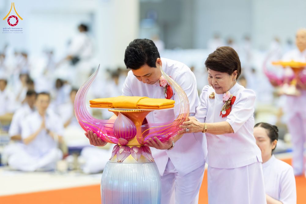 ชายและหญิงสวมชุดขาวกำลังประคองพานพุ่มดอกบัวที่มีผ้าไตรจีวรวางอยู่ด้านบนในพิธีทางศาสนา