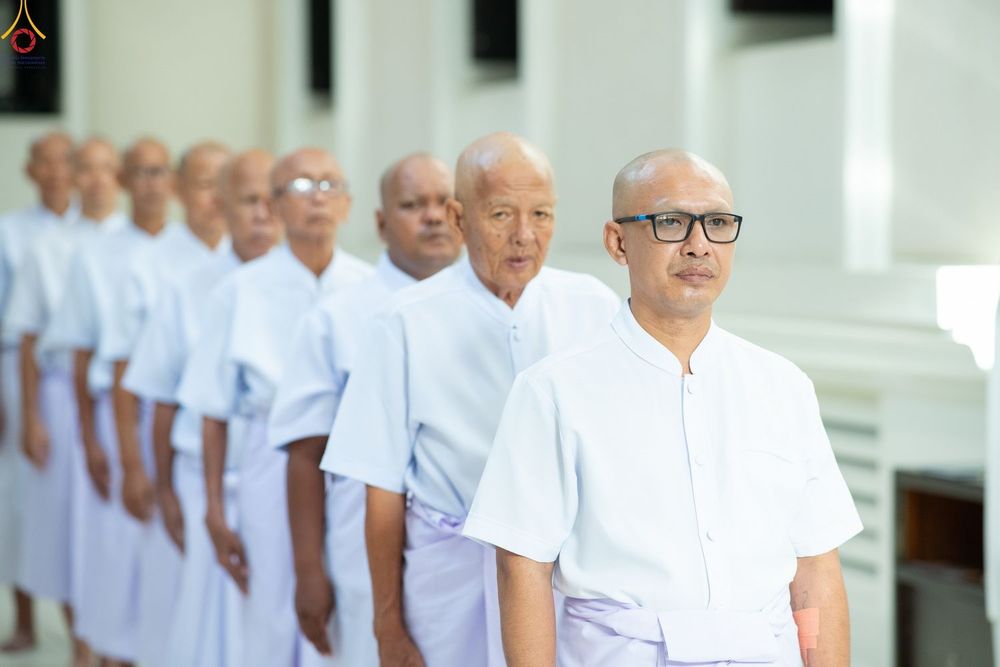 กลุ่มชายไทยโกนศีรษะสวมชุดสีขาวเรียงแถวอย่างเป็นระเบียบภายในอาคาร