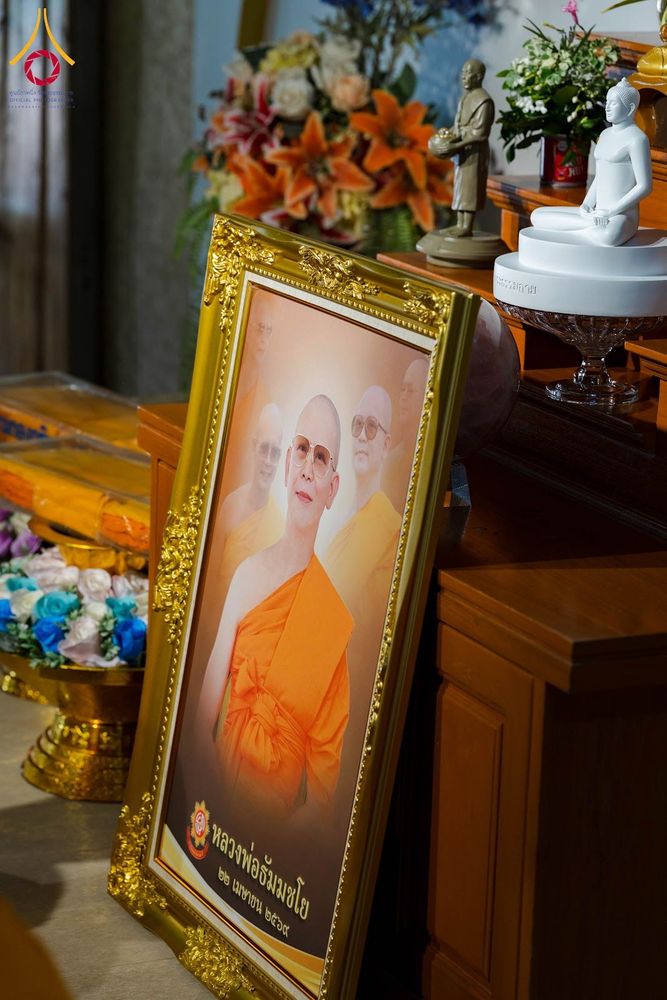 กรอบรูปสีทองบรรจุภาพถ่ายหลวงพ่อธัมมชโยตั้งอยู่บนโต๊ะไม้ข้างดอกไม้และพระพุทธรูป