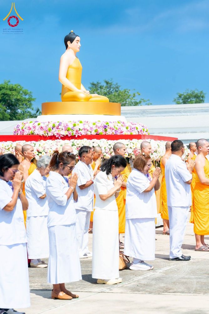 กลุ่มพุทธศาสนิกชนสวมชุดขาวและพระภิกษุสงฆ์กำลังยืนพนมมือประกอบพิธีหน้าพระพุทธรูปองค์สีทองที่ประดับด้วยดอกไม้