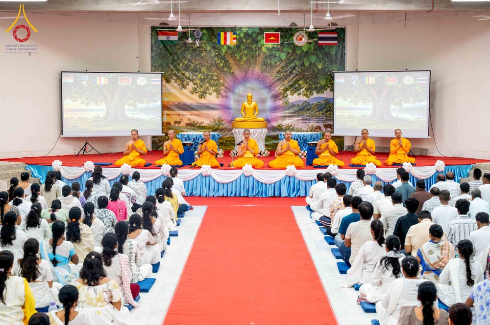 พระภิกษุสงฆ์และพุทธศาสนิกชนชุดขาวกำลังร่วมกิจกรรมปฏิบัติธรรมภายในหอประชุมที่มีฉากหลังเป็นภาพพระพุทธรูป