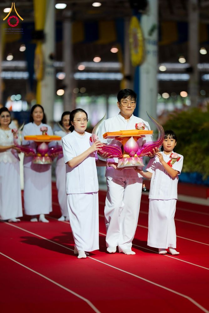 กลุ่มคนสวมชุดขาวถือพานพุ่มผ้าไตรเดินบนพรมสีแดงในพิธีทางศาสนา