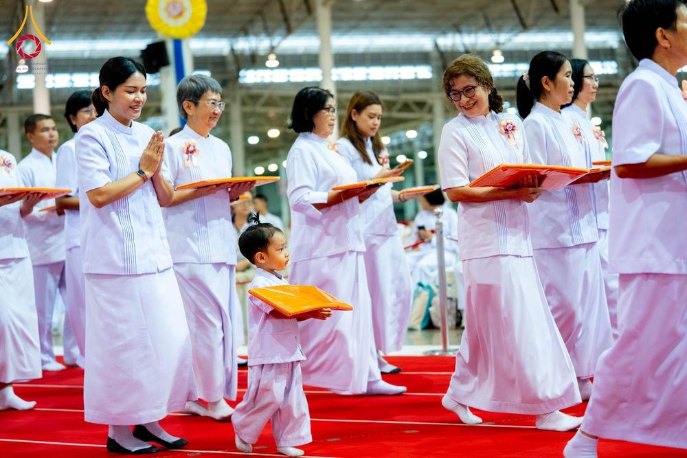 กลุ่มคนสวมชุดสีขาวเดินในขบวนพิธีการภายในอาคารกว้างขวาง โดยมีเด็กเล็กเดินนำหน้าและทุกคนถือซองสีส้ม