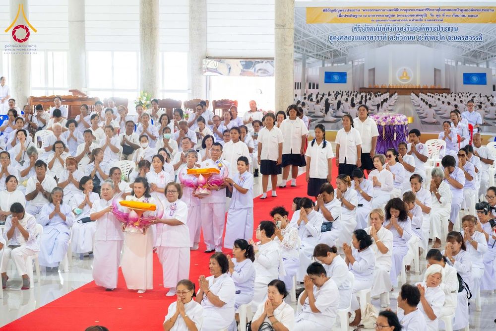 กลุ่มพุทธศาสนิกชนในชุดสีขาวกำลังร่วมพิธีทางศาสนาภายในอาคาร โดยมีผู้ถือพานพุ่มดอกบัวเดินนำขบวนบนพรมแดง