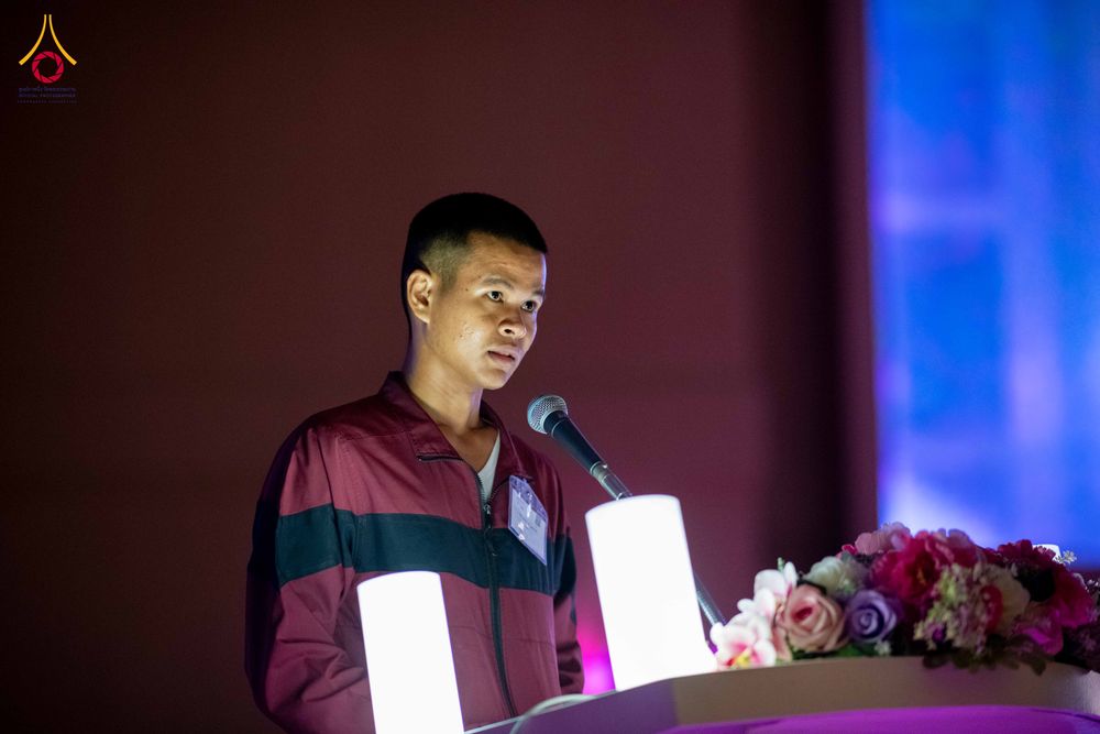 ชายหนุ่มสวมเสื้อแจ็กเก็ตสีแดงเข้มยืนพูดผ่านไมโครโฟนบนเวทีที่มีดอกไม้ประดับ