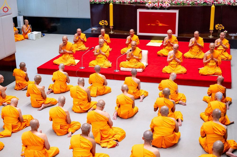 พระภิกษุและสามเณรจำนวนมากในจีวรสีเหลืองทองกำลังประกอบพิธีบรรพชาอุปสมบทภายในอาคารที่มีการจัดเตรียมสถานที่อย่างเป็นระเบียบ