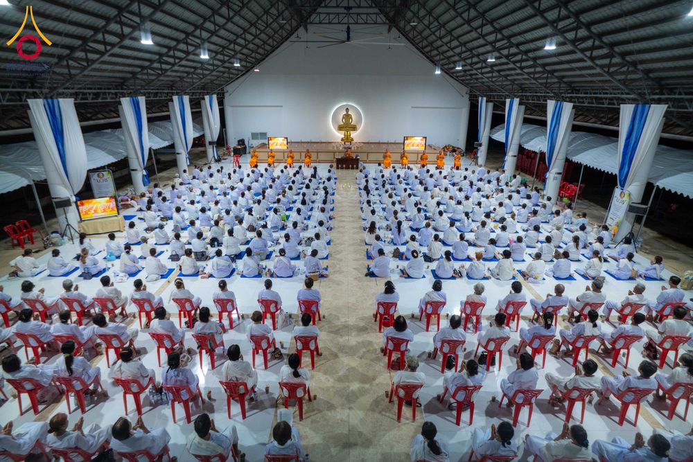 ผู้เข้าร่วมโครงการอบรมปฏิบัติธรรมจำนวนมากนั่งเรียงแถวในอาคารปฏิบัติธรรม โดยมีพระภิกษุสงฆ์นั่งอยู่ด้านหน้าใกล้พระพุทธรูป