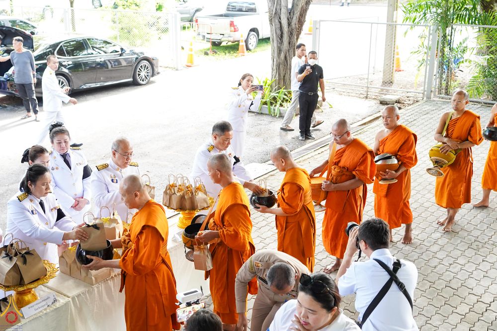 พระภิกษุสงฆ์ในจีวรสีส้มกำลังรับถวายเครื่องไทยธรรมจากเจ้าหน้าที่ในชุดเครื่องแบบสีขาวและผู้เข้าร่วมพิธี
