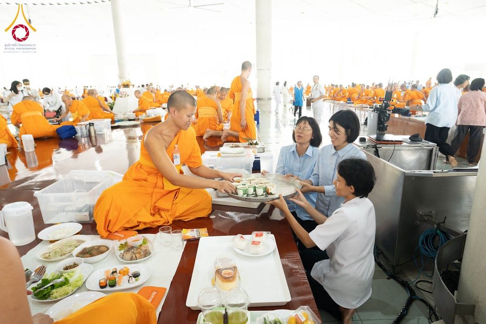 พระภิกษุสงฆ์กำลังรับถาดภัตตาหารจากสาธุชนภายในอาคารหอฉันที่มีพระภิกษุและผู้คนนั่งอยู่เป็นจำนวนมาก