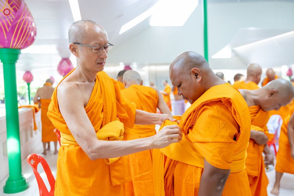 พระภิกษุสวมแว่นตากำลังช่วยจัดจีวรสีเหลืองทองให้แก่ผู้เข้าร่วมพิธีอุปสมบทหมู่