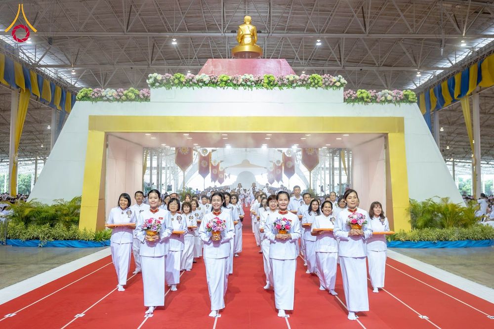 กลุ่มคนสวมชุดขาวถือพานดอกไม้เดินแถวบนพรมแดงในอาคารขนาดใหญ่