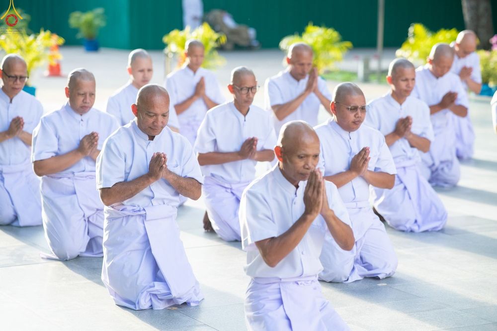 กลุ่มชายสวมชุดสีขาวนั่งคุกเข่าพนมมือเรียงแถวกันอย่างเป็นระเบียบในพิธีทางศาสนา