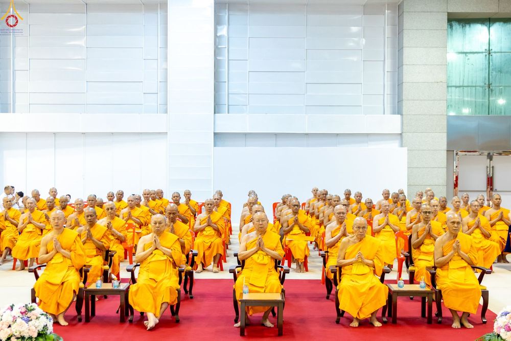 คณะสงฆ์จำนวนมากในจีวรสีเหลืองทองนั่งพนมมือบนเก้าอี้ภายในอาคารสภาธรรมกายสากล