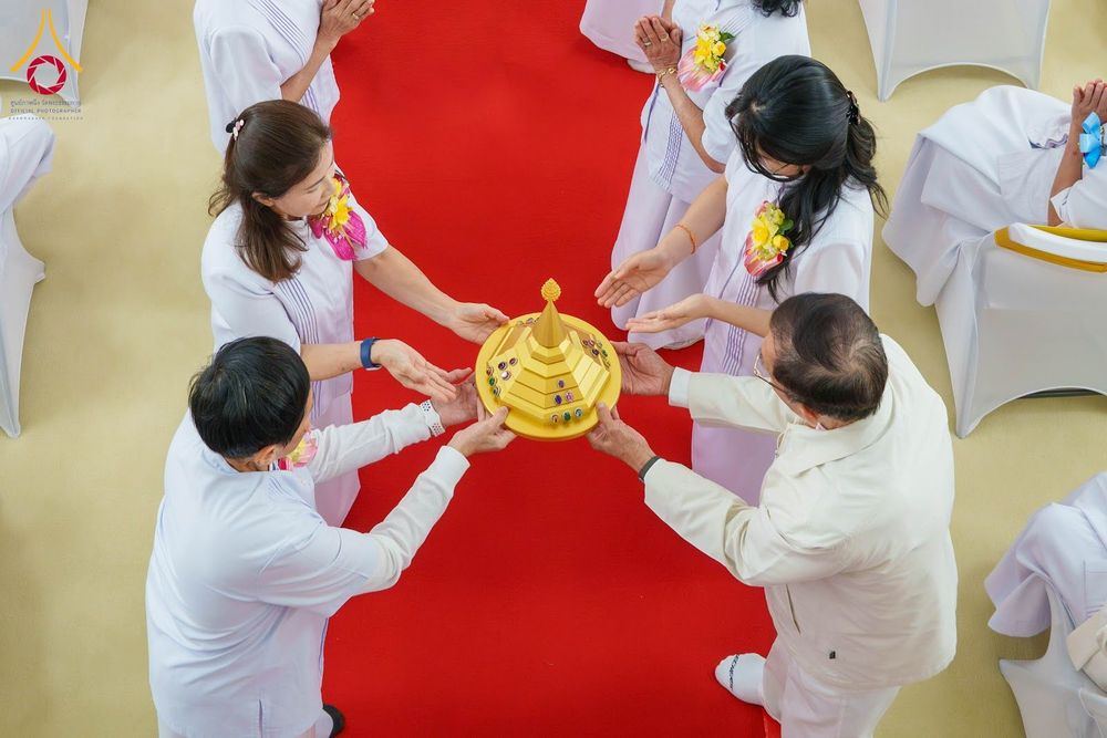 กลุ่มคนในชุดขาวกำลังช่วยกันถือพานพุ่มสีทองประดับอัญมณีเหนือพรมสีแดง