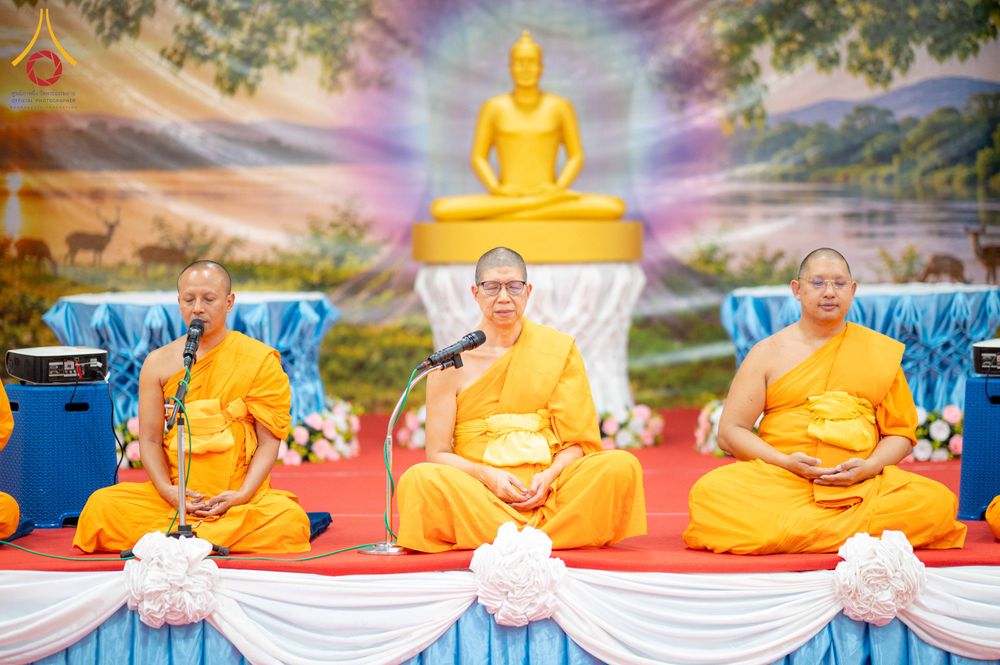 พระสงฆ์สามรูปนั่งสมาธิบนเวทีที่มีฉากหลังเป็นภาพพระพุทธรูปและธรรมชาติ