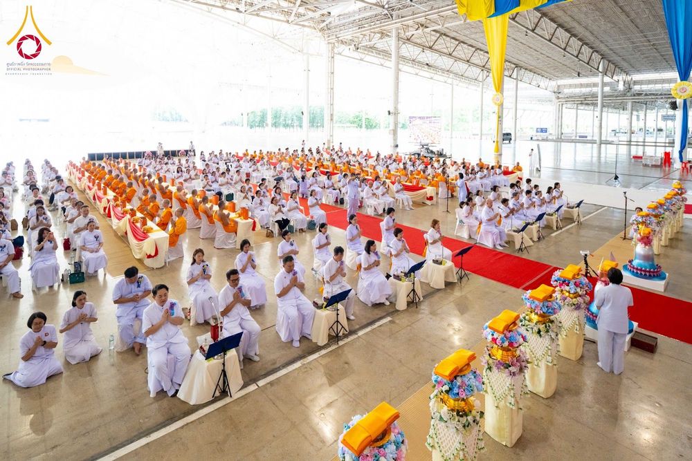 พุทธศาสนิกชนชุดขาวนั่งร่วมพิธีทางศาสนาภายในอาคารขนาดใหญ่ที่มีพระภิกษุสงฆ์และเครื่องไทยธรรม