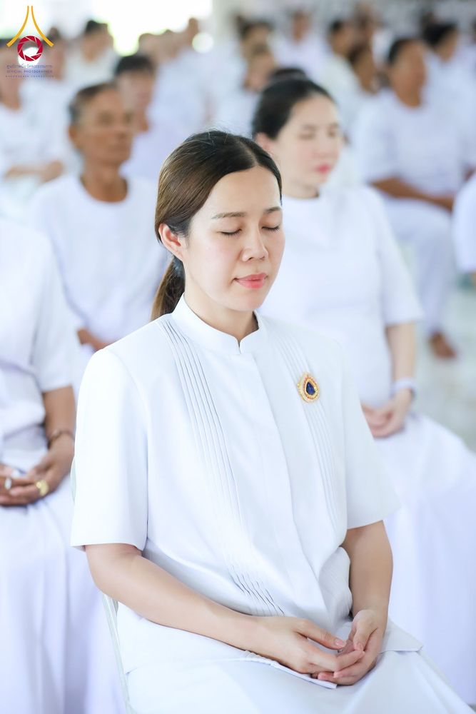 หญิงสาวสวมชุดสีขาวนั่งหลับตาทำสมาธิร่วมกับกลุ่มคนในชุดสีขาวภายในอาคาร
