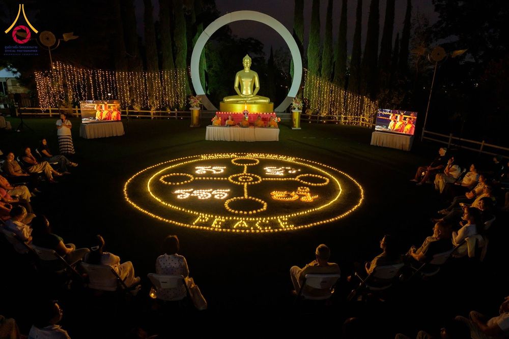 ผู้คนนั่งล้อมวงรอบการจุดประทีปเป็นรูปสัญลักษณ์และคำว่า PEACE หน้าพระพุทธรูปสีทองในยามค่ำคืน