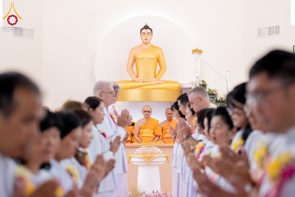 กลุ่มพุทธศาสนิกชนสวมชุดสีขาวกำลังพนมมือไหว้พระประธานสีทองและพระภิกษุสงฆ์ในพิธีทางศาสนา