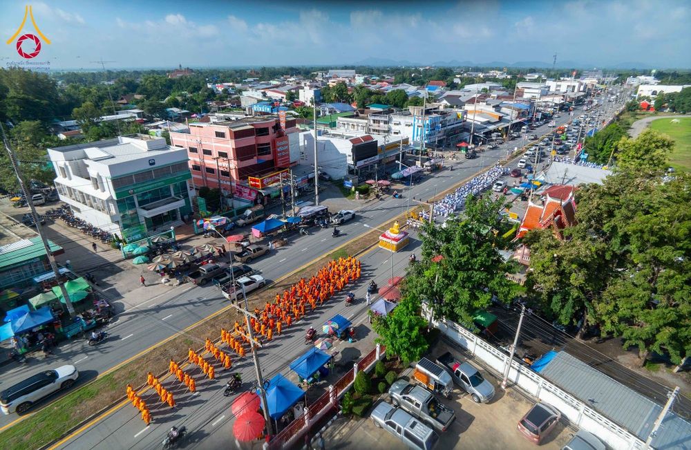 ขบวนพระภิกษุสงฆ์ห่มจีวรสีส้มเดินเรียงแถวบนถนนในเขตชุมชน โดยมีรถบุษบกอัญเชิญพระพุทธรูปนำหน้าขบวน