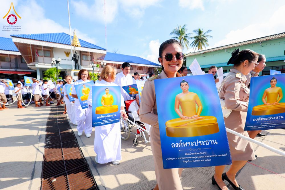 ผู้คนในชุดสุภาพและชุดขาวถือป้ายภาพพระประธานสีทองในขบวนแห่กลางแจ้ง