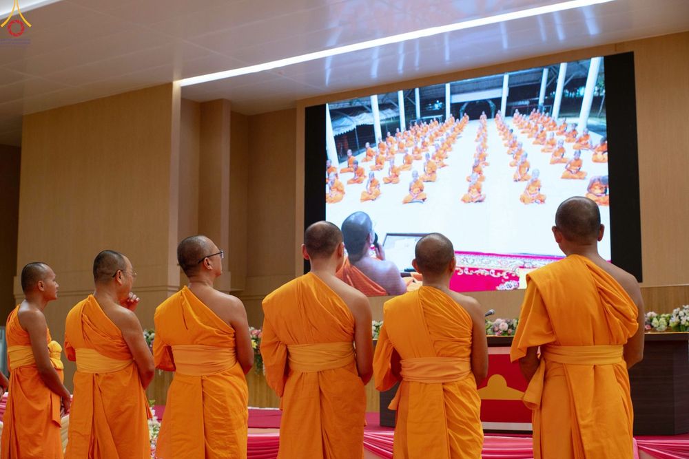 พระภิกษุสงฆ์หลายรูปยืนหันหลังมองหน้าจอขนาดใหญ่ที่แสดงภาพการปฏิบัติธรรมของพระภิกษุและสามเณรจำนวนมาก