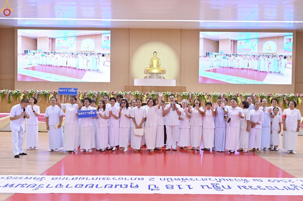 กลุ่มคนสวมชุดสีขาวจำนวนมากยืนเรียงแถวบนเวทีในห้องประชุมขนาดใหญ่