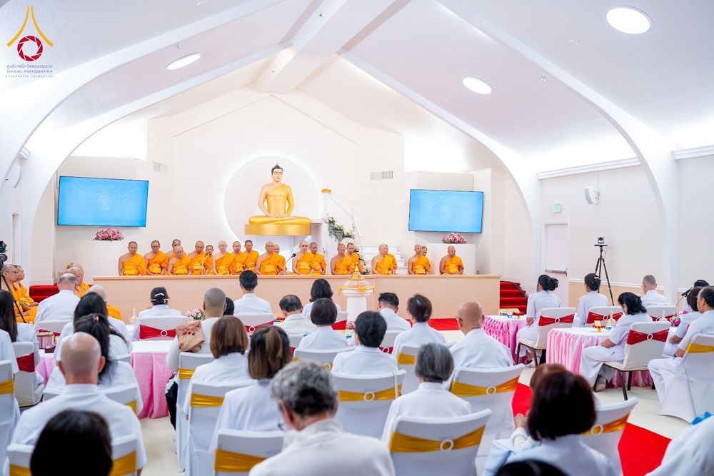 คณะสงฆ์และสาธุชนในชุดขาวร่วมพิธีทางศาสนาภายในอาคารที่มีพระพุทธรูปประธานเป็นศูนย์กลาง