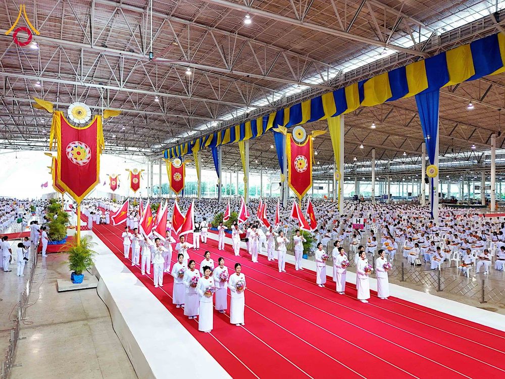 กลุ่มคนในชุดขาวเดินแถวบนพรมแดงในอาคารขนาดใหญ่ที่มีผู้คนนั่งร่วมพิธีจำนวนมาก