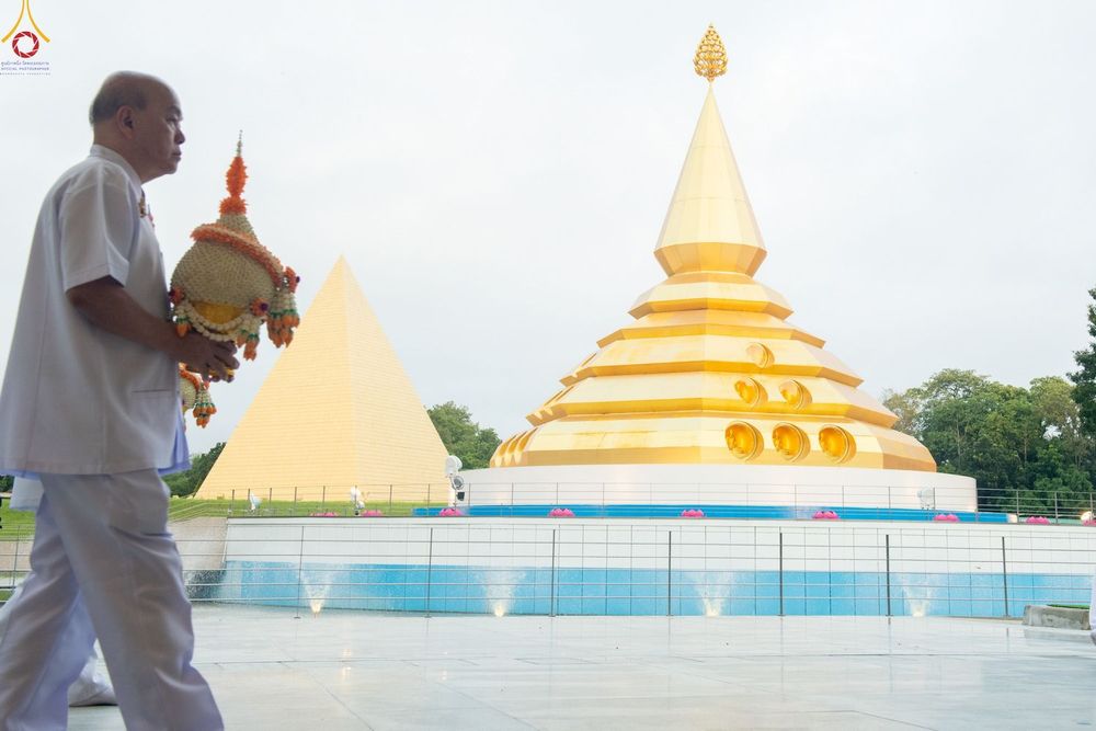 ชายสวมชุดสีขาวถือพานดอกไม้เดินผ่านเจดีย์สีทองและอาคารทรงพีระมิดสีทองในวัด