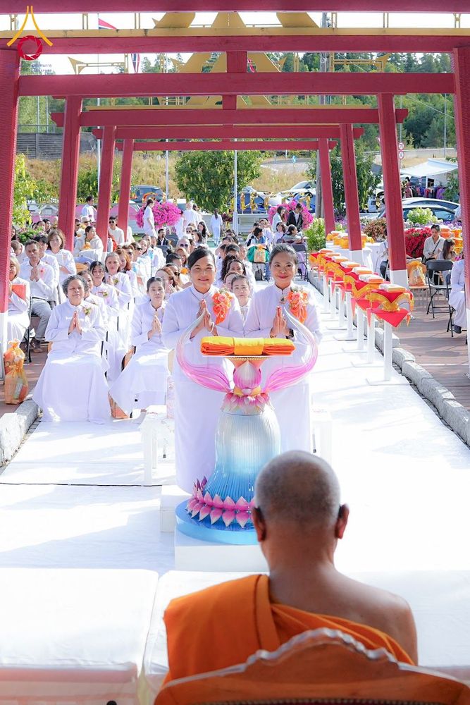 กลุ่มพุทธศาสนิกชนในชุดขาวกำลังประกอบพิธีทางศาสนา โดยมีตัวแทนถือพานผ้าไตรยืนอยู่ด้านหน้าแถวผู้เข้าร่วมพิธี