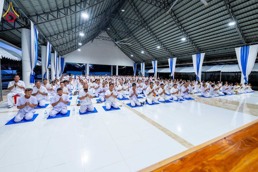 ผู้คนจำนวนมากสวมชุดขาวนั่งพนมมือบนเบาะรองนั่งภายในอาคารปฏิบัติธรรม