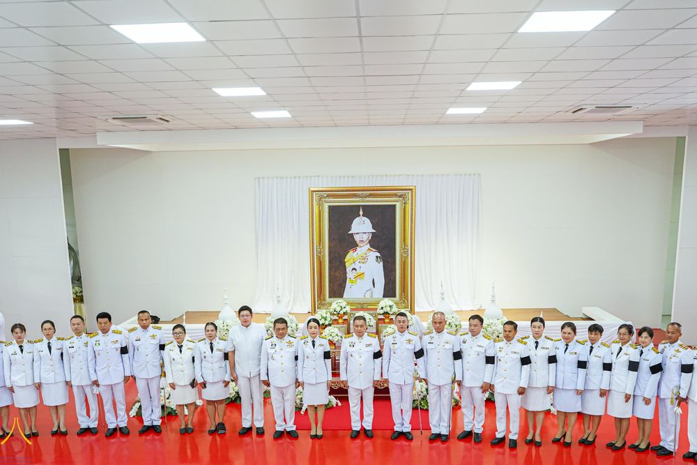 กลุ่มบุคคลในชุดเครื่องแบบปกติขาว ยืนเรียงแถวหน้าพระฉายาลักษณ์ภายในอาคาร