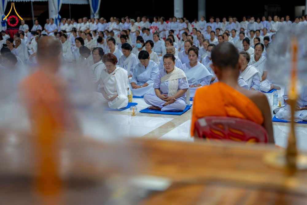 กลุ่มคนสวมชุดขาวนั่งสมาธิบนพื้นภายในอาคารโดยมีพระภิกษุนั่งนำอยู่ด้านหน้า