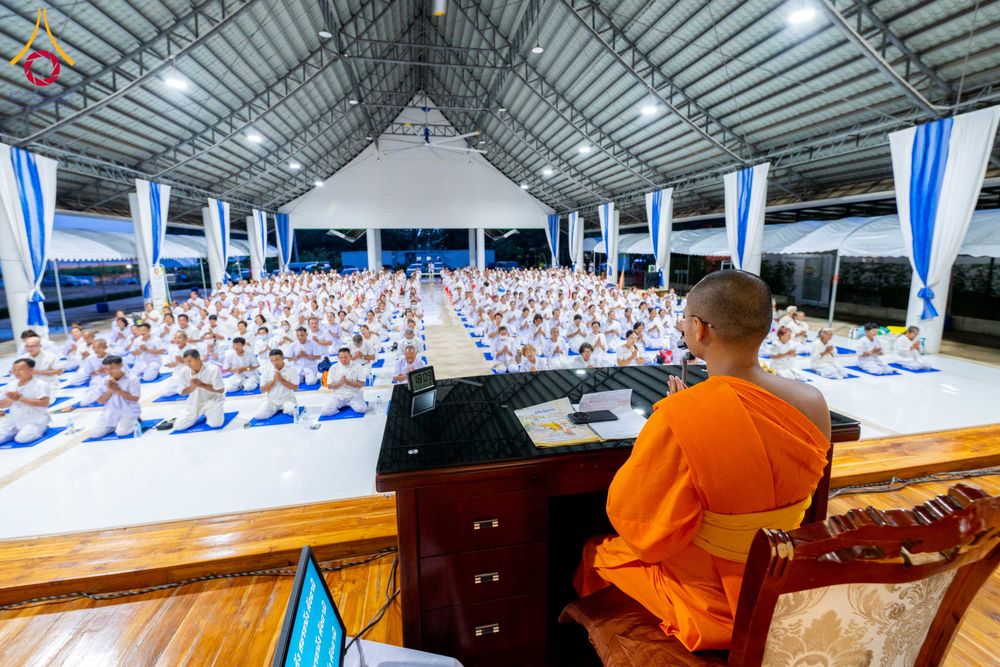 พระภิกษุสงฆ์นั่งบนเวทีหันหลังให้กล้องกำลังบรรยายธรรมแก่ผู้เข้าร่วมโครงการที่นั่งเรียงแถวปฏิบัติธรรมภายในอาคาร