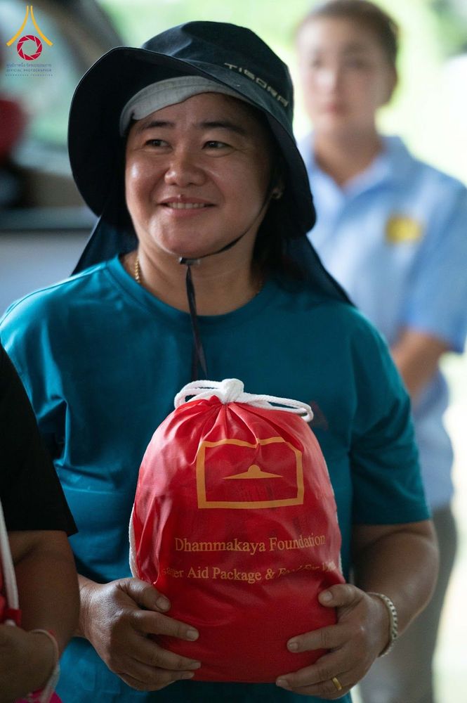 หญิงไทยสวมหมวกถือถุงยังชีพสีแดงของมูลนิธิธรรมกายด้วยรอยยิ้ม