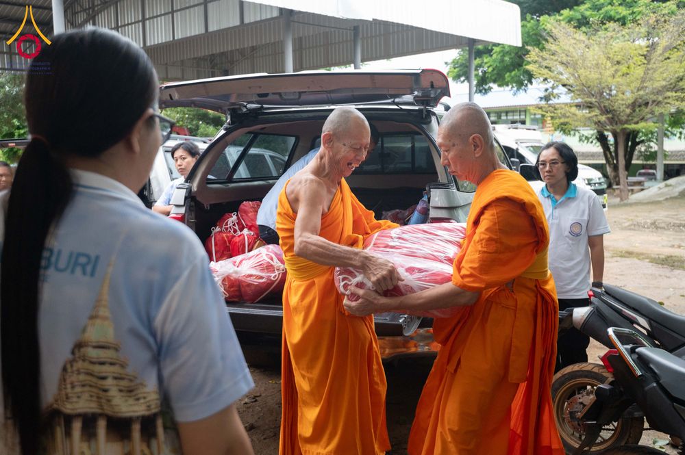 พระสงฆ์สองรูปกำลังช่วยกันยกถุงสิ่งของบรรเทาทุกข์สีแดงออกจากท้ายรถยนต์