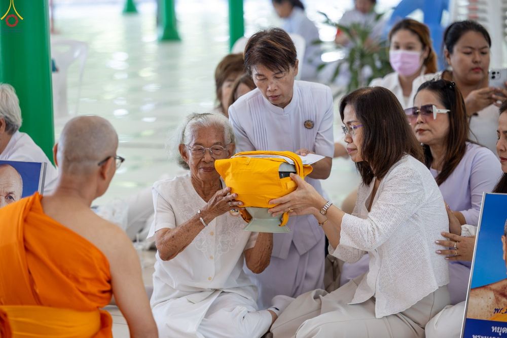 หญิงสูงวัยและผู้หญิงสวมชุดขาวกำลังประคองบาตรสีเหลืองเพื่อถวายแด่พระภิกษุสงฆ์ในพิธีทางศาสนา