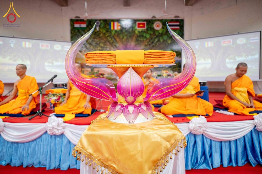 พานพุ่มดอกบัววางผ้าไตรจีวรสีเหลืองทอง โดยมีพระภิกษุสงฆ์นั่งสมาธิอยู่บนเวทีด้านหลัง