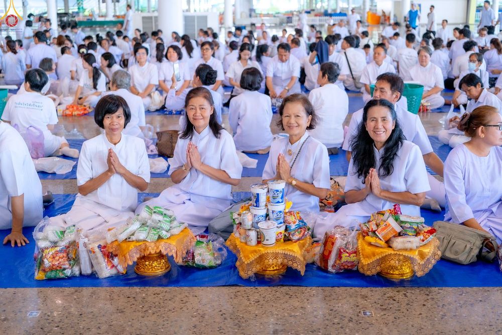 กลุ่มสตรีสี่ท่านสวมชุดขาวนั่งพนมมือเตรียมตักบาตรพร้อมพานอาหารแห้ง โดยมีผู้คนจำนวนมากนั่งอยู่ด้านหลังภายในอาคาร