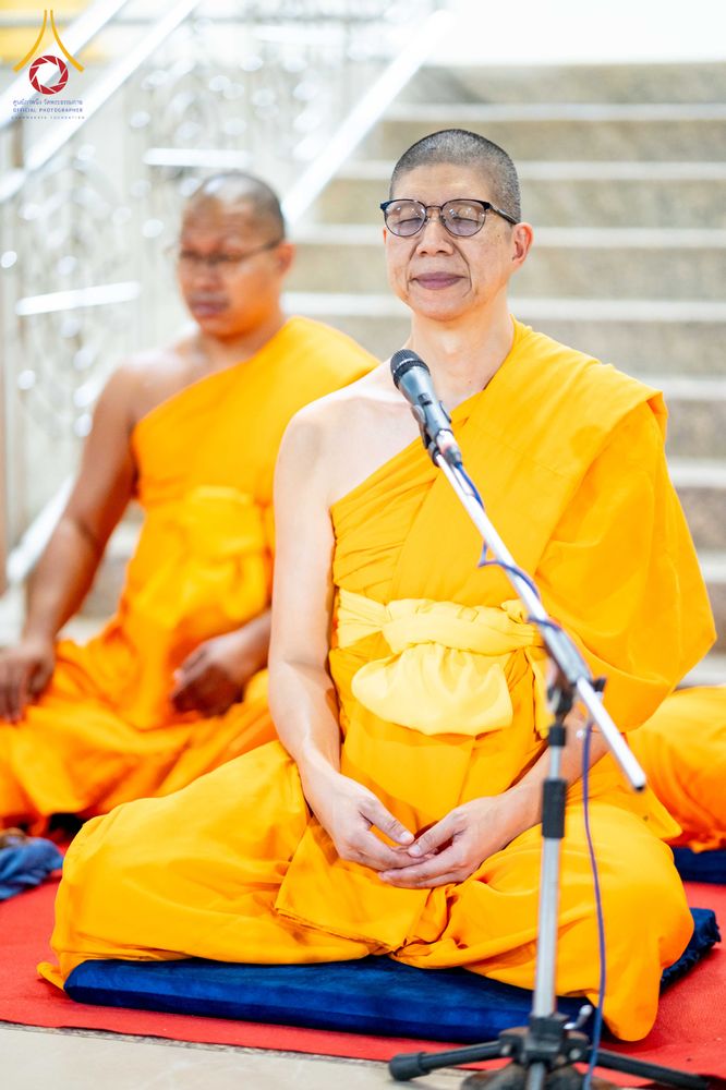 พระภิกษุสงฆ์สวมแว่นตานั่งสมาธิและพูดผ่านไมโครโฟนในชุดจีวรสีเหลือง