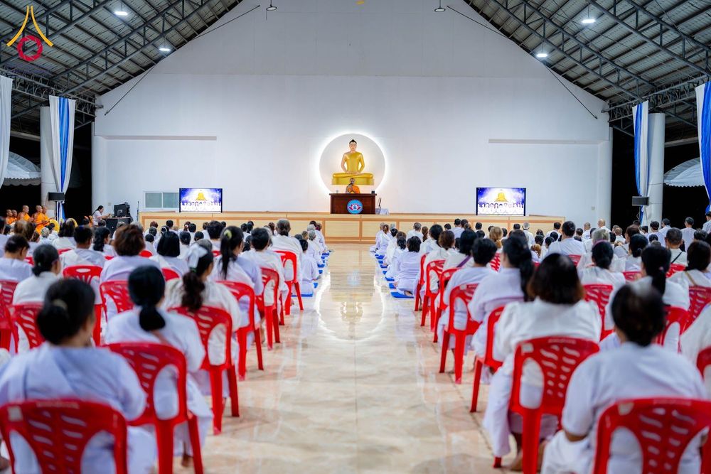 กลุ่มคนสวมชุดขาวนั่งฟังธรรมภายในอาคารที่มีพระพุทธรูปบนเวที