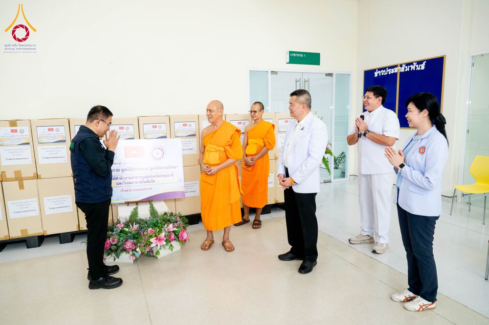 พระภิกษุและเจ้าหน้าที่ยืนร่วมในพิธีมอบกล่องยาและเวชภัณฑ์จำนวนมากเพื่อสาธารณสงเคราะห์ภายในอาคาร