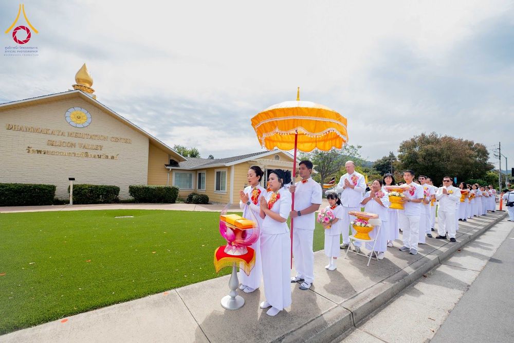 กลุ่มคนชุดขาวถือพานผ้าป่าและร่มสีเหลืองเดินแถวหน้าอาคารวัดพระธรรมกายซิลิคอนวัลเลย์