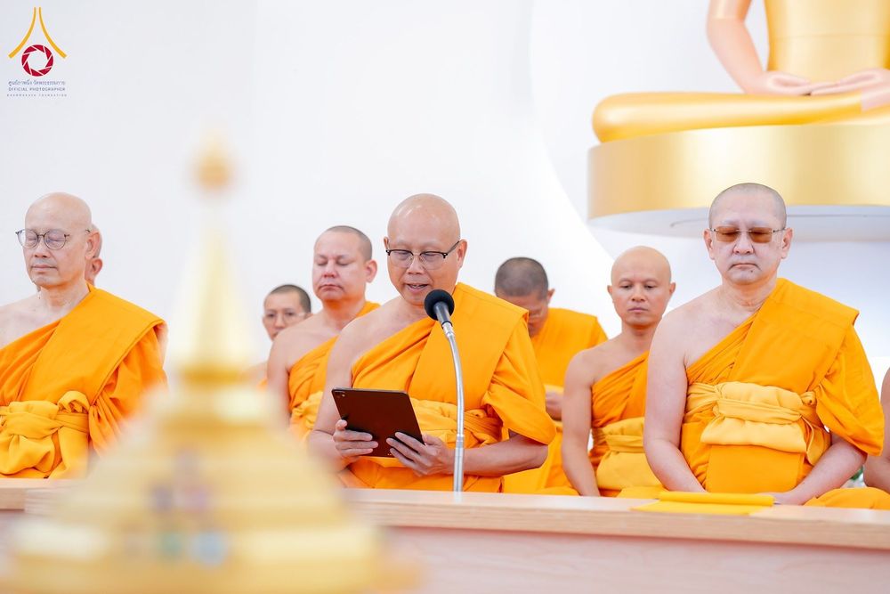 คณะสงฆ์หลายรูปนั่งเรียงแถวประกอบพิธีกรรมทางศาสนา โดยมีพระภิกษุรูปหนึ่งกำลังอ่านบทสวดจากแท็บเล็ตผ่านไมโครโฟน