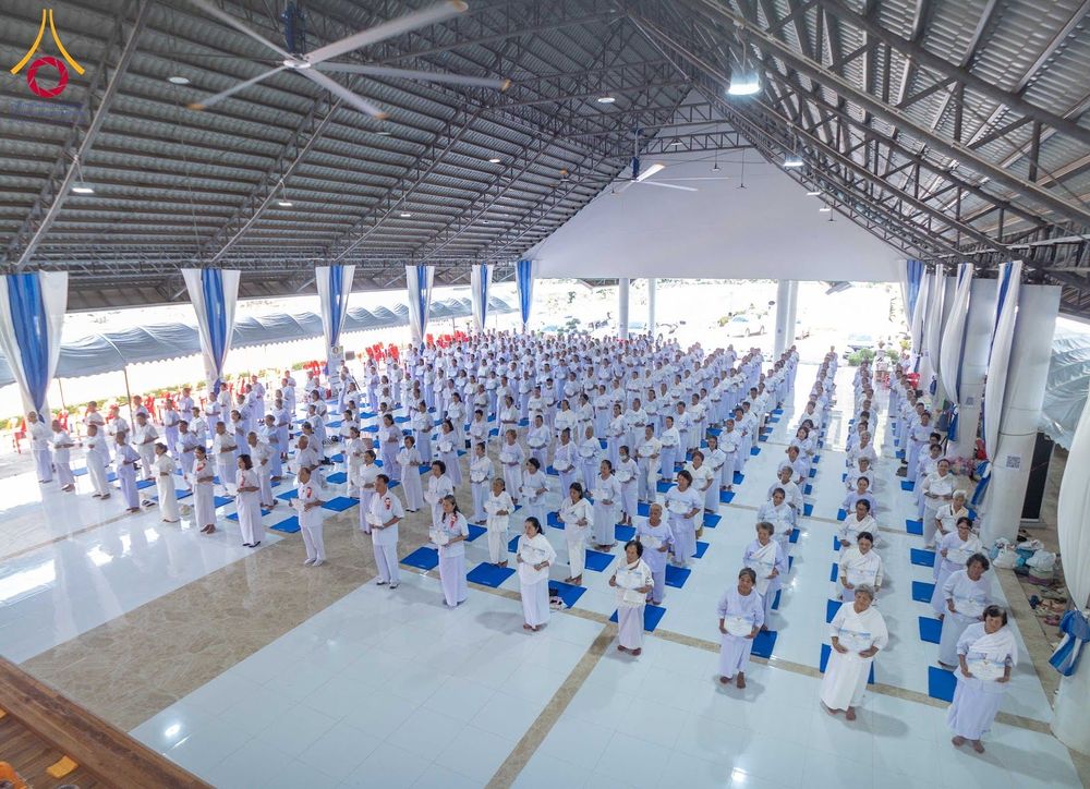 กลุ่มคนสวมชุดสีขาวจำนวนมากยืนเรียงแถวปฏิบัติธรรมภายในอาคาร