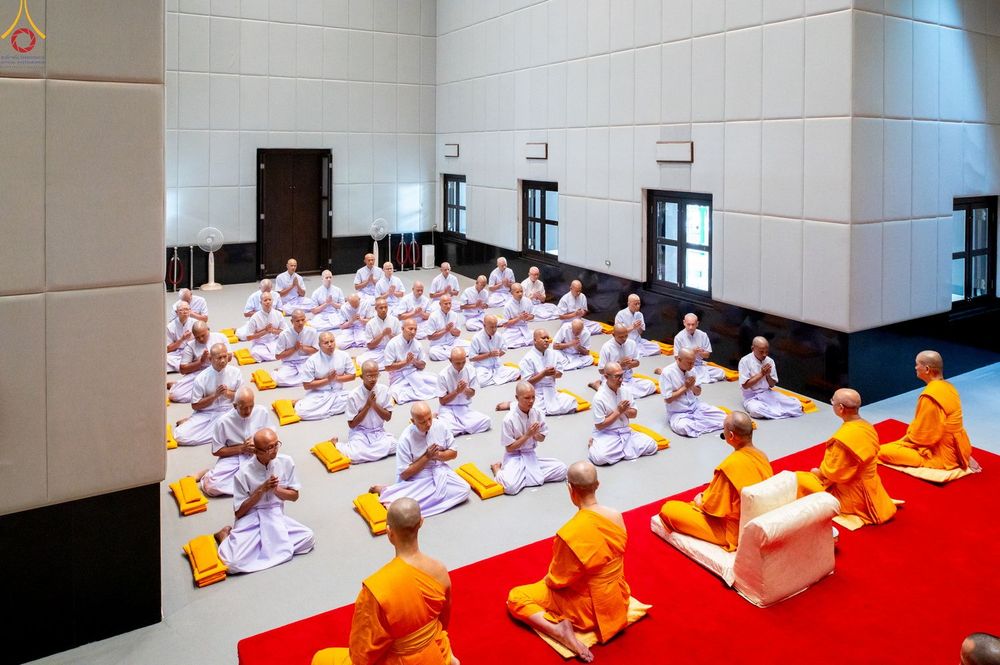กลุ่มผู้เข้าร่วมพิธีบรรพชาอุปสมบทในชุดสีขาวนั่งพนมมือเรียงแถวภายในอาคาร โดยมีพระภิกษุในชุดจีวรสีส้มทำหน้าที่เป็นประธานสงฆ์นั่งอยู่ด้านหน้า